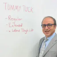 Dr. Rodriguez standing in front of a white board where he's written, Tummy Tuck with regular, extended, and with lateral thigh lift listed below that