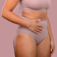 Woman wearing a nude compression garment after tummy tuck and Brazilian butt lift surgery, gently holding her abdomen.